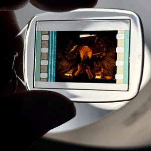 May include: A silver-colored keychain with a rectangular film strip insert. The film strip shows a scene with a figure in a dark setting. The film strip has blue, white, and tan accents. The keychain is held by a person.