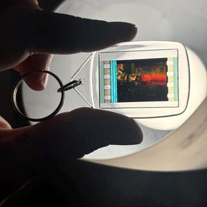 May include: A clear, rectangular keychain with a metal ring. The keychain holds a small, colorful image of a film strip. The film strip shows a scene with red and yellow hues. The keychain is held up against a bright light.