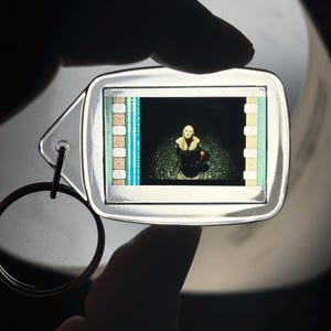 May include: Silver-toned keychain with a rectangular insert featuring a film strip design. The film strip displays a scene with a person in a dark setting. The keychain has a circular ring for attaching keys.