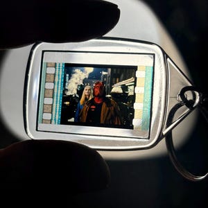 May include: A silver-colored keychain with a rectangular film strip insert featuring a scene from a movie. The film shows a red-skinned character with horns and a woman with blonde hair. The keychain has a silver ring attached.
