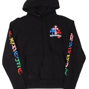 Op de afbeelding: Zwarte hoodie met een trekkoord en een voorzak. De mouwen hebben kleurrijke letters en de borst toont een kruisontwerp in roze, blauw en rood. De tekst "CHROME HEARTS" staat eronder.