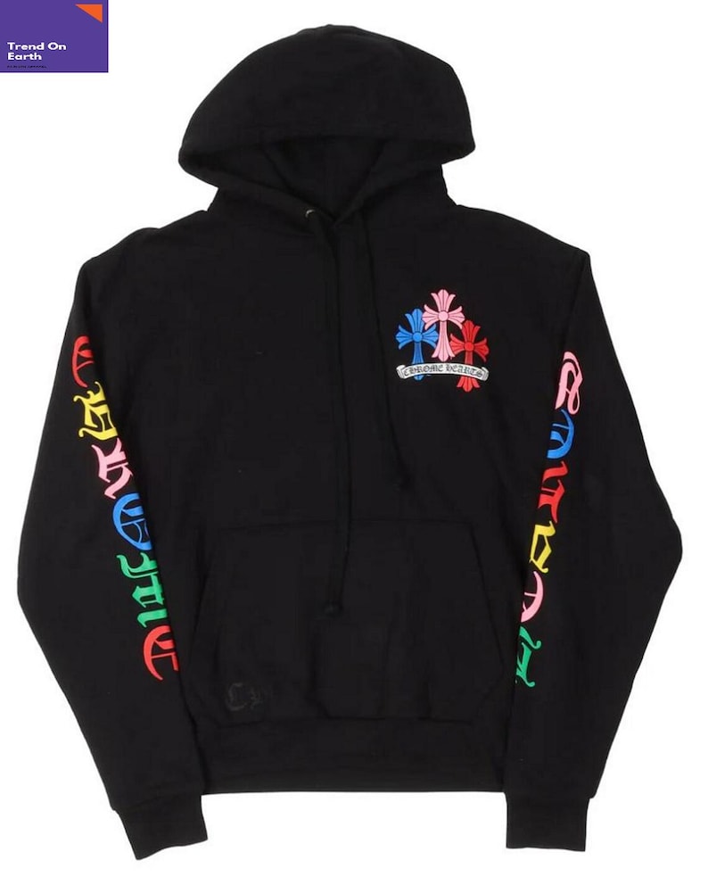 Op de afbeelding: Zwarte hoodie met een trekkoord en een voorzak. De mouwen hebben kleurrijke letters en de borst toont een kruisontwerp in roze, blauw en rood. De tekst "CHROME HEARTS" staat eronder.