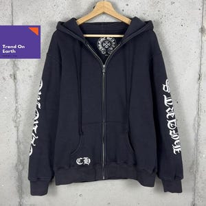 Può includere: Una felpa nera con zip e logo bianco in stile gotico sul petto e sulle maniche. L'etichetta interna sul collo mostra un emblema circolare Chrome Hearts. La parte anteriore presenta una cerniera, un cappuccio con coulisse e due tasche.
