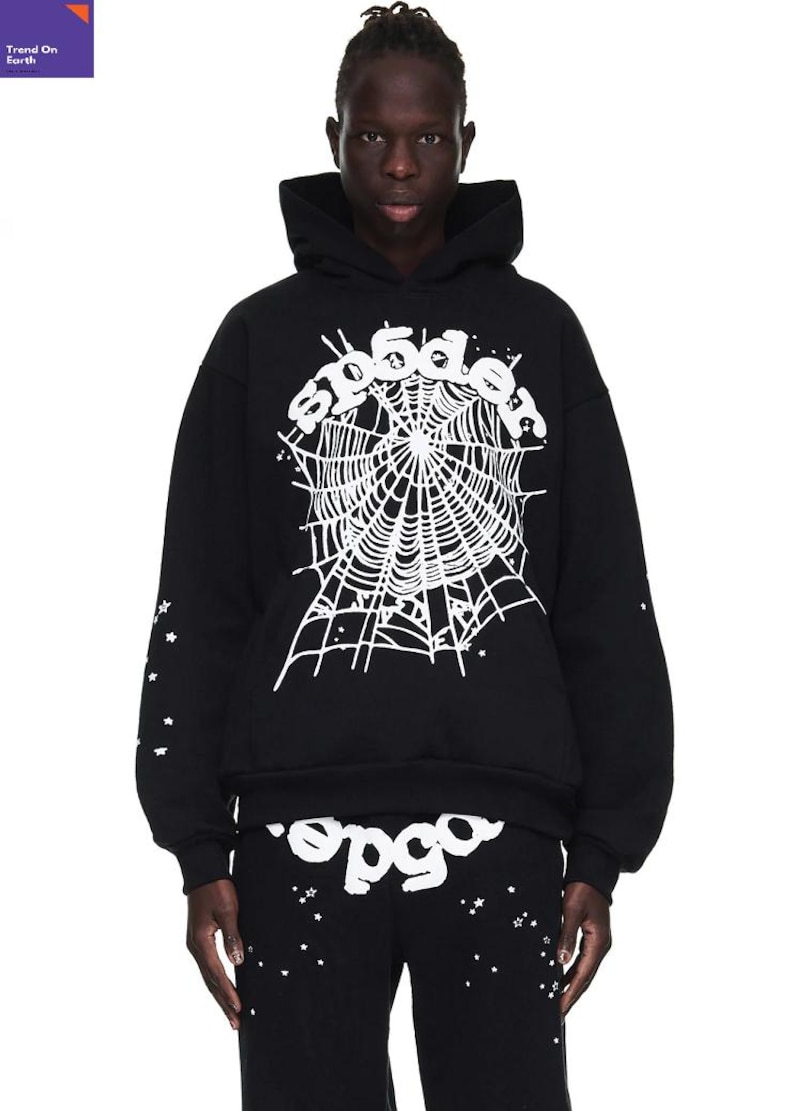 以下が含まれることがあります： 白いクモの巣のグラフィックと「spider」の文字が入った黒いパーカーとパンツ。袖とパンツには白い星が散りばめられています。ストリートウェアコレクションの一部です。