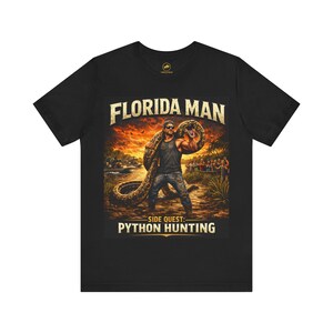 May include: Black t-shirt featuring a graphic of a man holding a large snake, with the text "FLORIDA MAN" above and "SIDE QUEST: PYTHON HUNTING" below. The design includes a sunset background and a crowd of people.