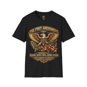 May include: Black t-shirt with a detailed graphic of an eagle holding an American flag and olive branch. The text reads "THE FIRST AMENDMENT" and "FREEDOM OF RELIGION SPEECH PRESS ASSEMBLY PETITION OUR AMERICAN RIGHTS."