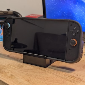 Pode incluir: Um console de jogos portátil preto com detalhes em azul e laranja está encaixado em um suporte preto. O console está em uma superfície de madeira, com um monitor e outros acessórios de jogos ao fundo. A tela é preta.