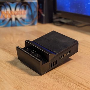 May include: A black electronic docking station featuring a rectangular base with a slanted top surface, a front-facing charging port, and two USB ports on the side. The device has a textured finish and includes vertical ventilation slots along the side.