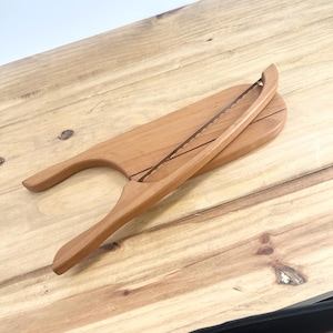 May include: A wooden bread knife with a serrated blade and a handle that is shaped like a bread loaf. The knife is resting on a wooden cutting board.