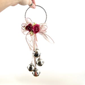 May include: A decorative silver metal ring with a twisted design, adorned with a pink bow, burgundy flowers, and dangling silver bells. The bells have star cutouts and hang from pink ribbons, creating a festive ornament.