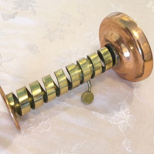 May include: A vintage brass and copper candle holder with a spiral design. The holder has a round base and a knob on the side.