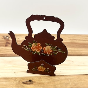 May include: A decorative, dark brown wooden teapot-shaped trivet with a matching small stand. The trivet features hand-painted orange and yellow floral designs with green leaves. The handle has a hole for hanging. The stand has a similar floral design.