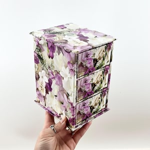 May include: A small white and purple floral patterned wooden box with three drawers.