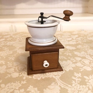 May include: A white ceramic coffee grinder with a wooden base and a drawer. The grinder has a brown trim around the top and bottom. The handle is made of wood.