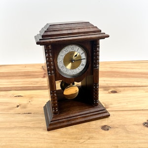 May include: A dark brown wooden mantel clock with a gold-colored clock face. The clock has Roman numerals and a pendulum. The clock is rectangular with a decorative top and base.