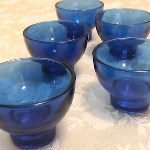 May include: Five cobalt blue glass bowls with a flared rim and a rounded base. The bowls are arranged in a row on a white lace tablecloth.
