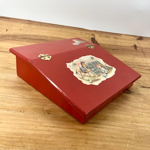 May include: A red wooden slanted desk box with two brass-colored metal hinges on the top edge. The lid features a central decal depicting a child on a rocking horse and a smaller decal near the top edge, both showing illustrations of children.