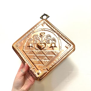 May include: A copper trivet with a raised design of a heart and flowers in a basket. The trivet has a decorative border and a hanging loop.