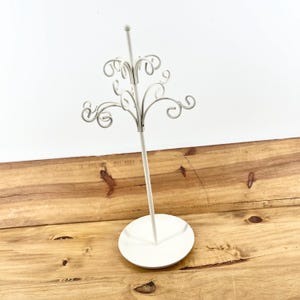 May include: A white metal jewelry display stand with a round base and a tall, slender stem. The top features decorative, swirling scrollwork for hanging earrings or necklaces. The stand is on a wooden surface.