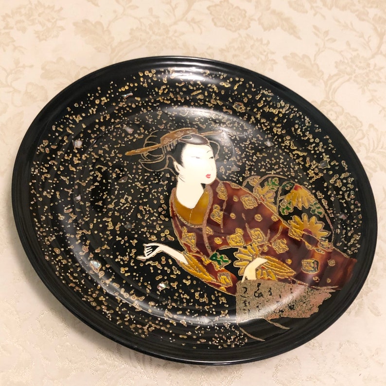 May include: A black lacquer plate with a gold and red design of a Japanese woman in a kimono. The plate has a speckled gold background.