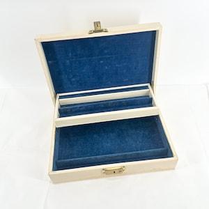 May include: A white jewelry box with a blue velvet interior and a gold latch. The box has a hinged lid and a compartment for storing jewelry.