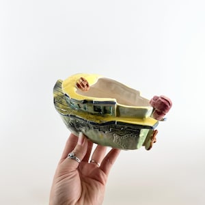 May include: A ceramic boat-shaped planter with a yellow rim and accents. The boat has green, blue, and brown details, with a red propeller and a small brown object on the top. The planter is held in a hand.