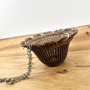 May include: A brown, vintage hanging basket with a decorative, scalloped rim. The basket is made of carved wood and features vertical slats. A metal chain is attached for hanging. The basket is empty and sits on a wooden surface.
