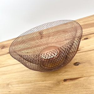 May include: A copper-colored wire mesh bowl with a rounded shape. The bowl has a textured, open weave design and sits on a wooden surface. The bowl has a smaller, solid copper-colored sphere at the base.