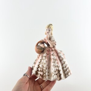 May include: A porcelain figurine of a person with blonde hair wearing a long, wide dress decorated with a textured pattern of small, raised white and pink flowers. The figure holds a small, brown, woven-style basket in one hand.