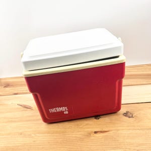 May include: A red and white Thermos 16 cooler with a white lid. The cooler has a rectangular shape with rounded corners and a handle. The word "THERMOS" and the number "16" are printed on the side of the cooler.