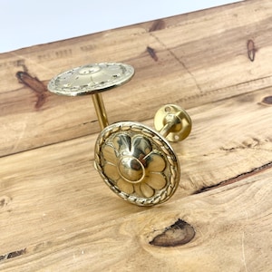 May include: Two brass-colored curtain tiebacks with a floral design. The tiebacks have a circular plate with a flower in the center, surrounded by a decorative border. The stems are straight and the mounting plates are small and round.