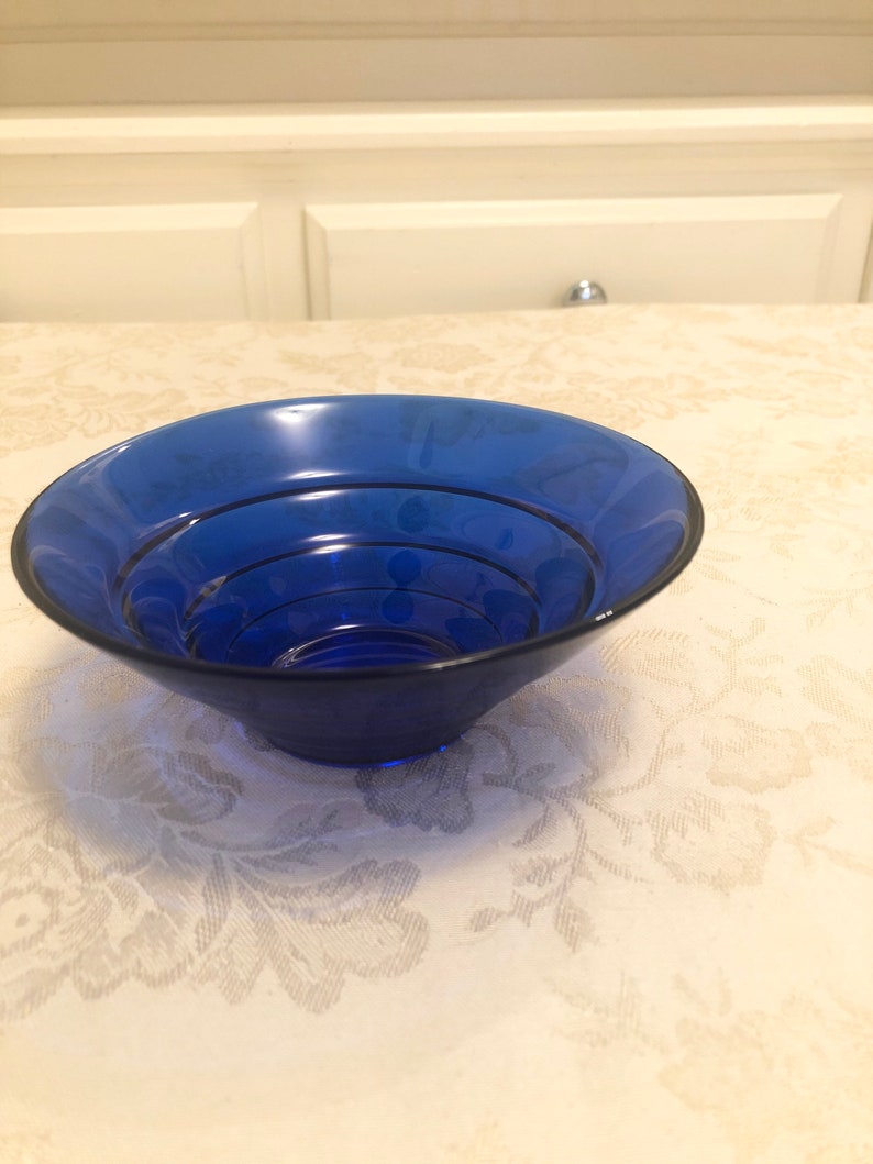 May include: A cobalt blue glass bowl with a fluted rim. The bowl is empty and sitting on a white tablecloth with a floral pattern.
