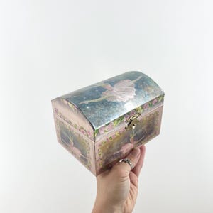 May include: A rectangular jewelry box with a curved lid, featuring a painted ballerina in a pink tutu on the top and front panels. The box is decorated with pink floral borders, a metallic latch, and a blue and gold background design.