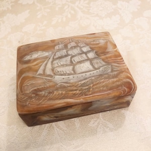 May include: A rectangular, brown and white, carved stone box with a ship design on the lid. The ship is carved in white and the waves are carved in brown.