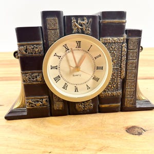 May include: A decorative clock shaped like a stack of books. The clock face is gold with black Roman numerals and reads "QUARTZ." The books are dark brown with gold accents. The clock is approximately 6 inches tall.