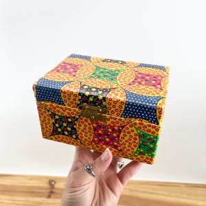 May include: A square, decorative box with a colorful floral pattern. The box features a yellow background with red, green, and blue floral designs. It has a brass-colored clasp and is held by a hand.