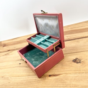 May include: A two-tiered pink jewelry box with a hinged lid and a green velvet interior. The top tier has several compartments for rings and earrings, while the bottom tier is a larger, open space.