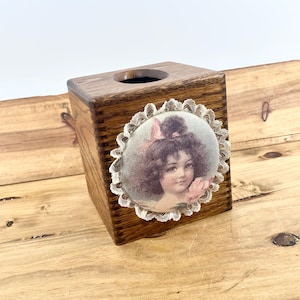 May include: A wooden tissue box with a vintage-style image of a girl with curly hair and a pink bow, framed by a white lace border. The box is made of dark brown wood and has a square shape.