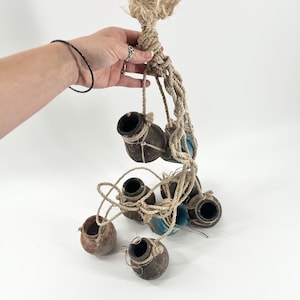 May include: A set of six small clay pots hanging from a woven rope. The pots are brown and have a rustic appearance. The rope is natural in color and has a knot at the top.