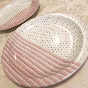 May include: Two pink and white ceramic plates with a polka dot and striped design. The plates are oval shaped and have a slightly raised rim.