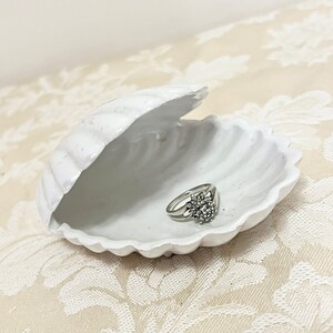 May include: A white ceramic seashell ring holder with a silver ring with a cluster of clear stones inside.