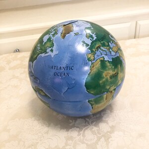 May include: A blue and green globe piggy bank with a slot on the top. The globe has a map of the world with the continents labeled in black text. The text "ATLANTIC OCEAN" is visible on the globe.