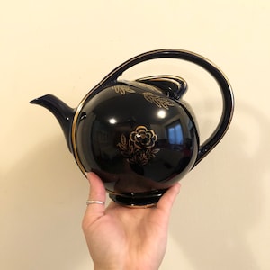 May include: A navy blue teapot with a gold floral design and trim. The teapot has a large, curved handle and a spout. The floral design includes a rose and leaf accents. The teapot is held by a hand.