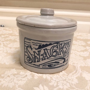 May include: A white ceramic jar with a blue and white design that says "SNACKS". The jar has a lid and is perfect for storing snacks.