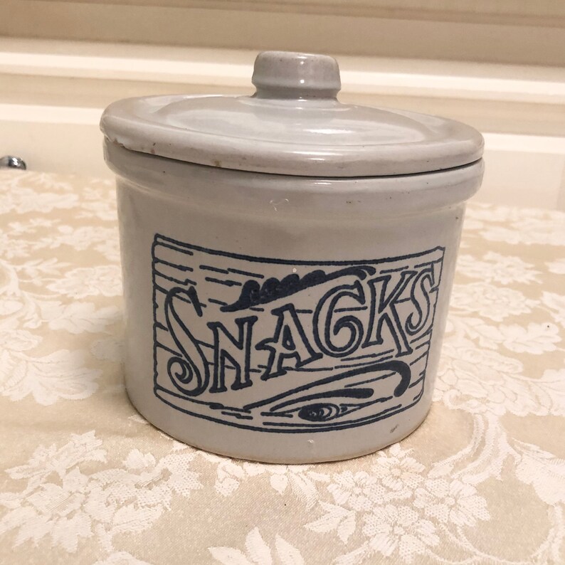 May include: A white ceramic jar with a blue and white design that says "SNACKS". The jar has a lid and is perfect for storing snacks.