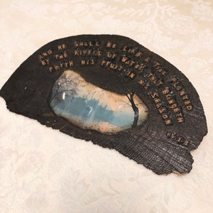 May include: A dark brown wooden plaque with a light brown inscription that reads "And he shall be like a tree planted by the rivers of water that bringeth forth his fruit in his season 1:3". The plaque has a small rectangular picture of a forest scene with a blue sky and white clouds.