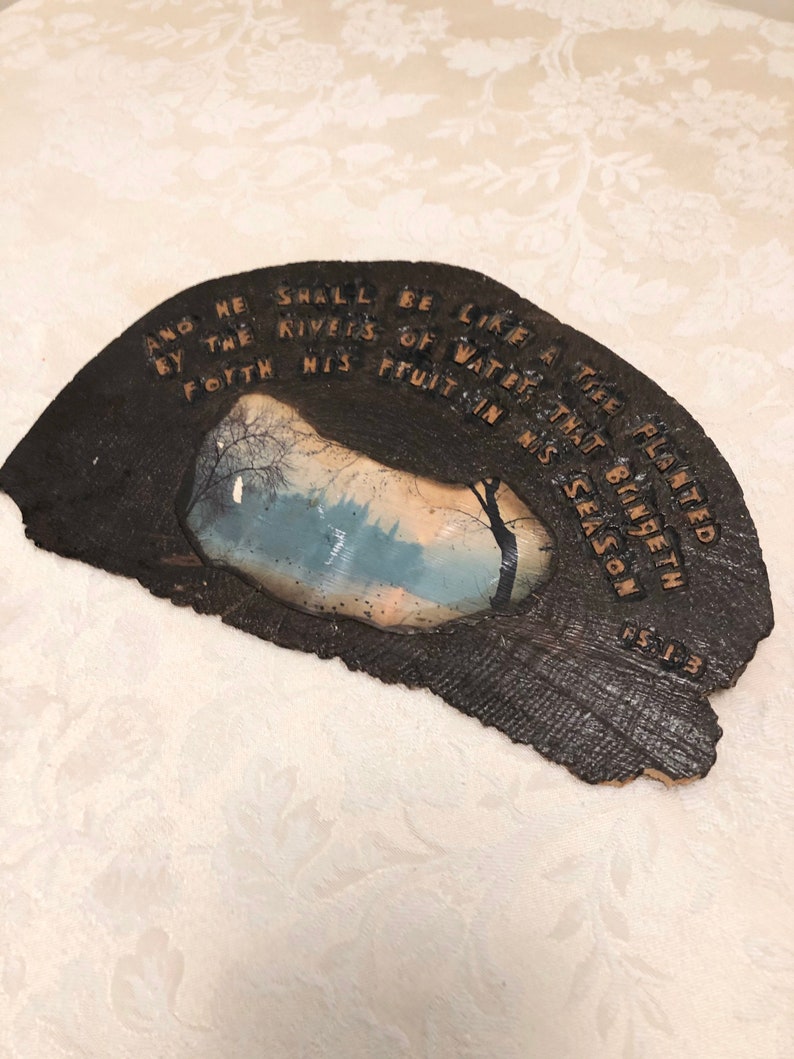 May include: A dark brown wooden plaque with a light brown inscription that reads "And he shall be like a tree planted by the rivers of water that bringeth forth his fruit in his season 1:3". The plaque has a small rectangular picture of a forest scene with a blue sky and white clouds.