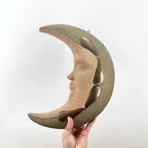 May include: A crescent moon wall hanging with a textured olive green exterior and a light tan face. The moon features brass-colored accents and a hook for hanging. The celestial-themed decor is a unique piece for any room.