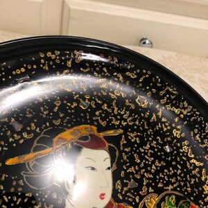 May include: A black lacquer plate with a gold and white design of a woman in traditional Japanese clothing. The plate is decorated with gold flecks.