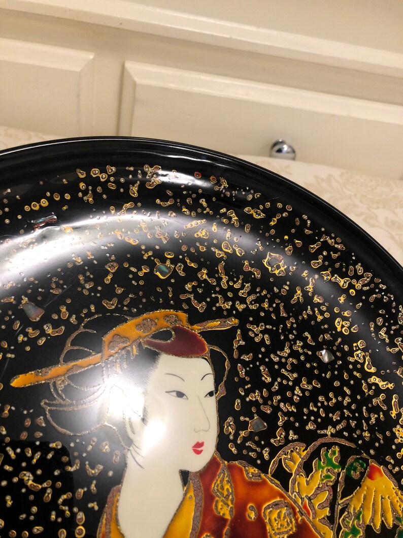 May include: A black lacquer plate with a gold and white design of a woman in traditional Japanese clothing. The plate is decorated with gold flecks.